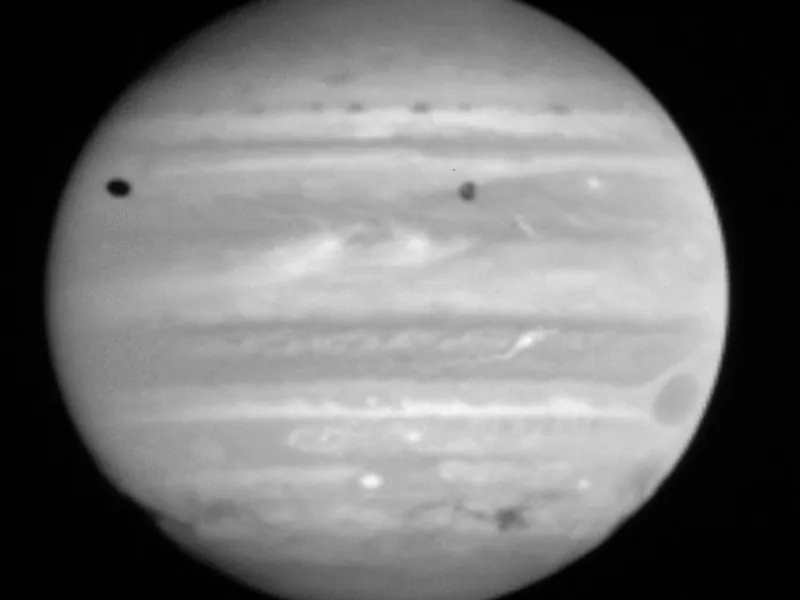 A grey picture of Jupiter with a row of dark spots across its upper half